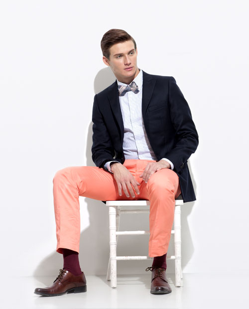 A seated man modeling a navy blazer, a blue pin-striped shirt, salmon pants and a bow tie.